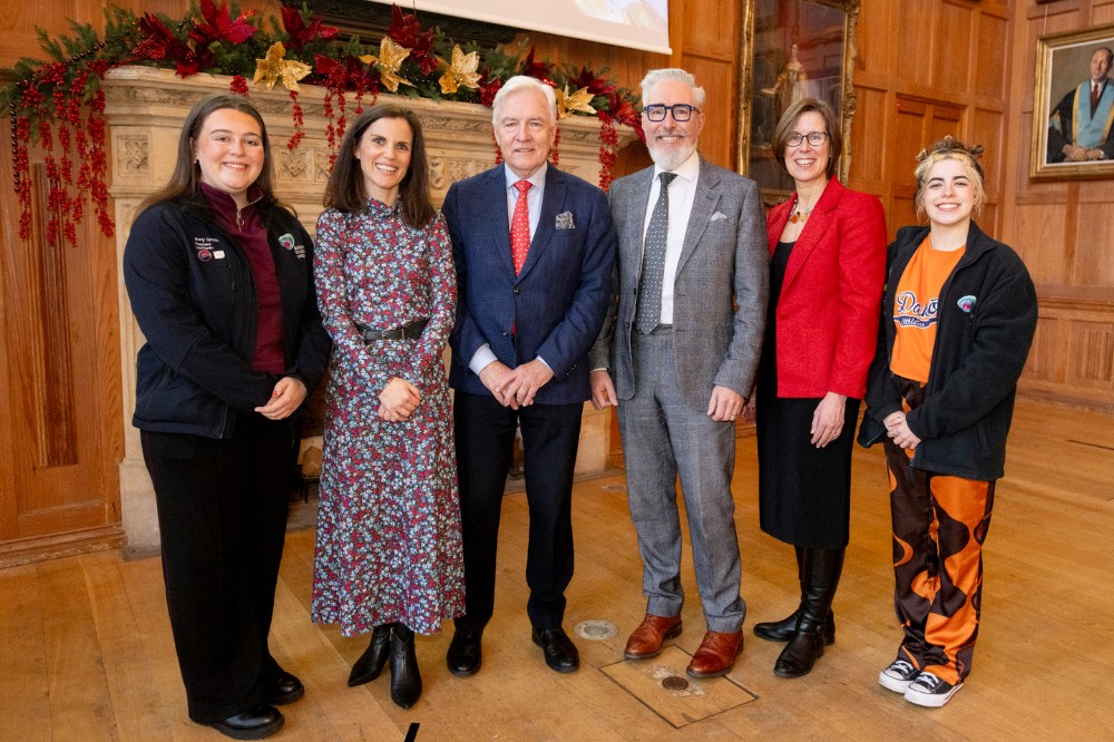 four women and two men in formal or business dress smiling to camera in front of a large, decorated, stone fireplace in an old indoor event space