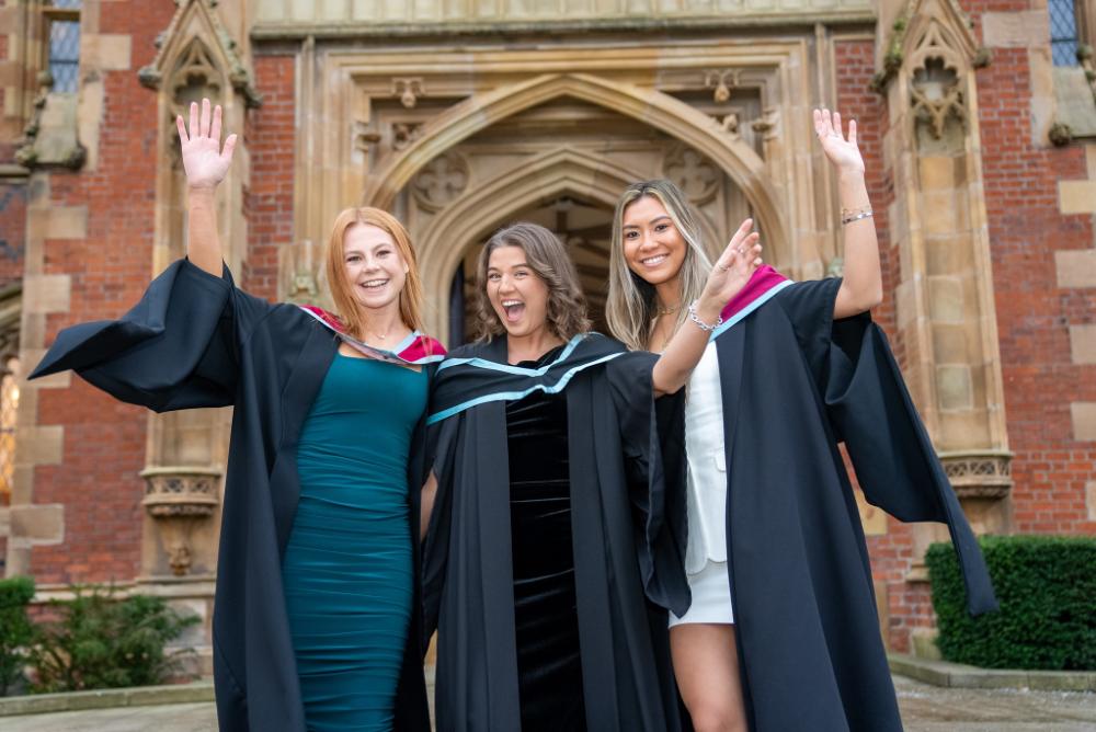 Three female graduates smiling and raising their hands in celebration, filled with joy outside Queen's University Belfast building.