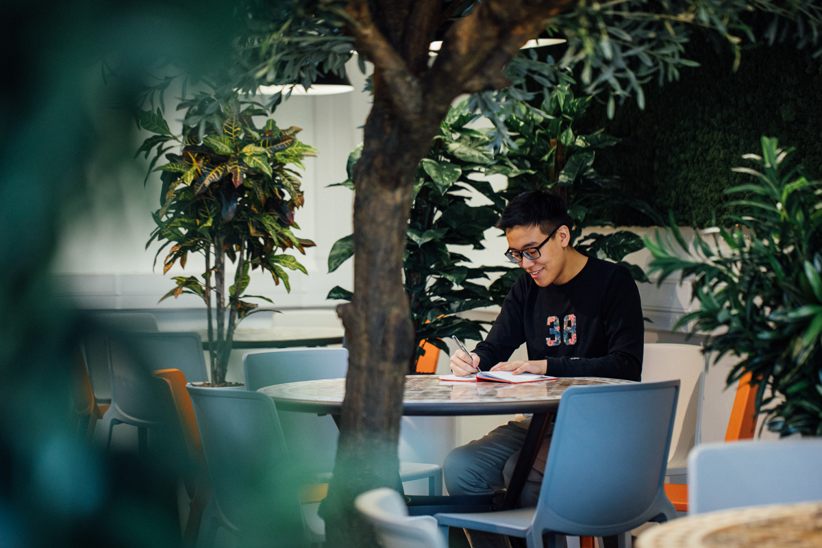 Image of student in Computer Science building with lots of plants, writing into a journal