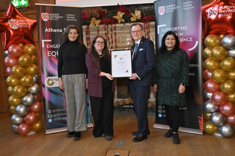 man and woman pose with a framed award certificate alongside two other women in an old event hall with large fireplace, pop-up stands and balloon pillars pictured to the sides and in the background
