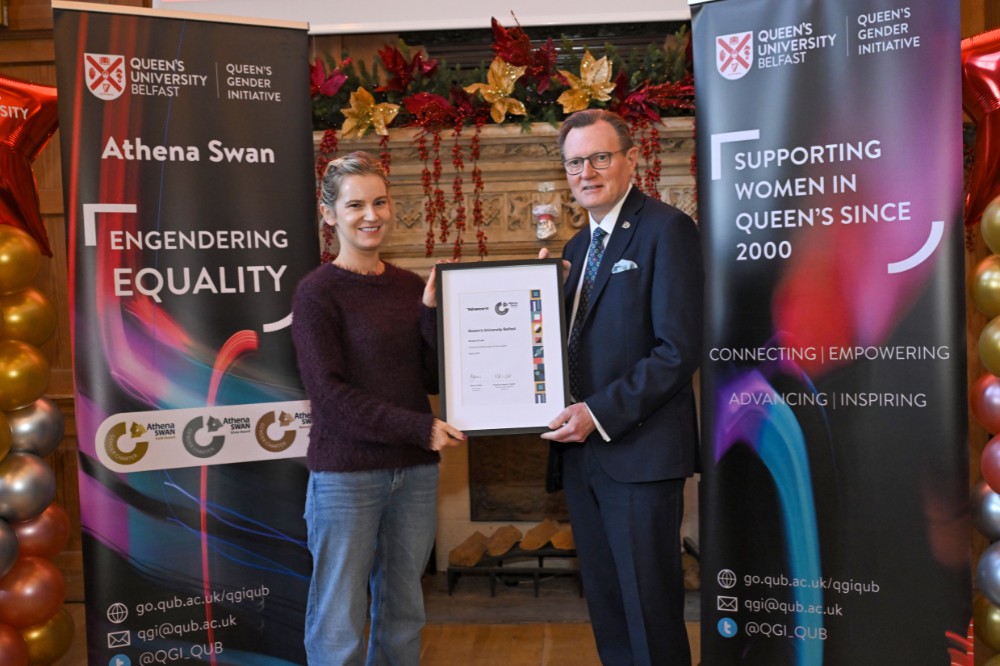 woman collecting a framed certificate award from a man in suit in an old wood-paneled event hall with pop-up stands and presentation screen in the background
