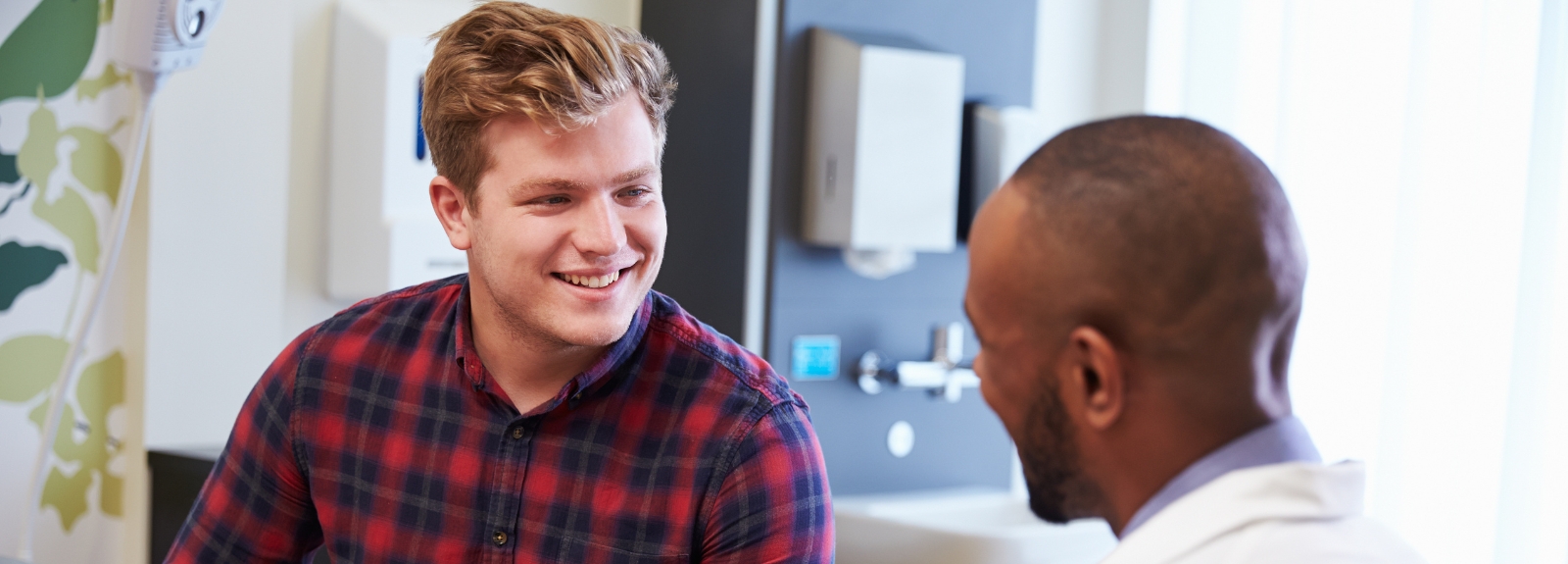 A young man wearing a red check shirt smiles while talking with a healthcare professional in a bright consultation room.