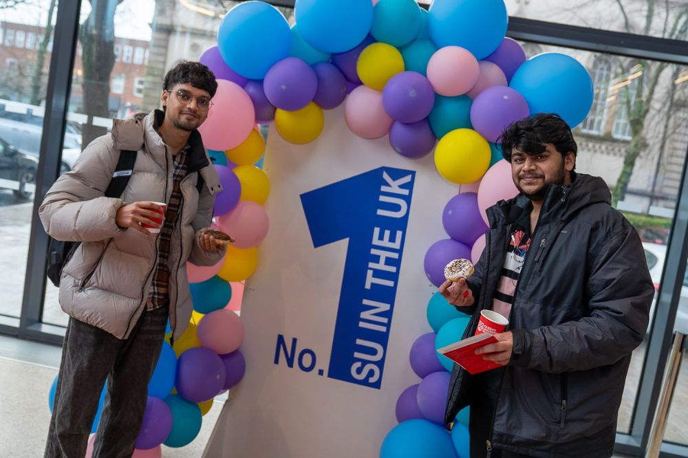 Two students stand by a balloon arch featuring a large “No. 1 SU in the UK” sign, holding doughnuts and hot drinks.