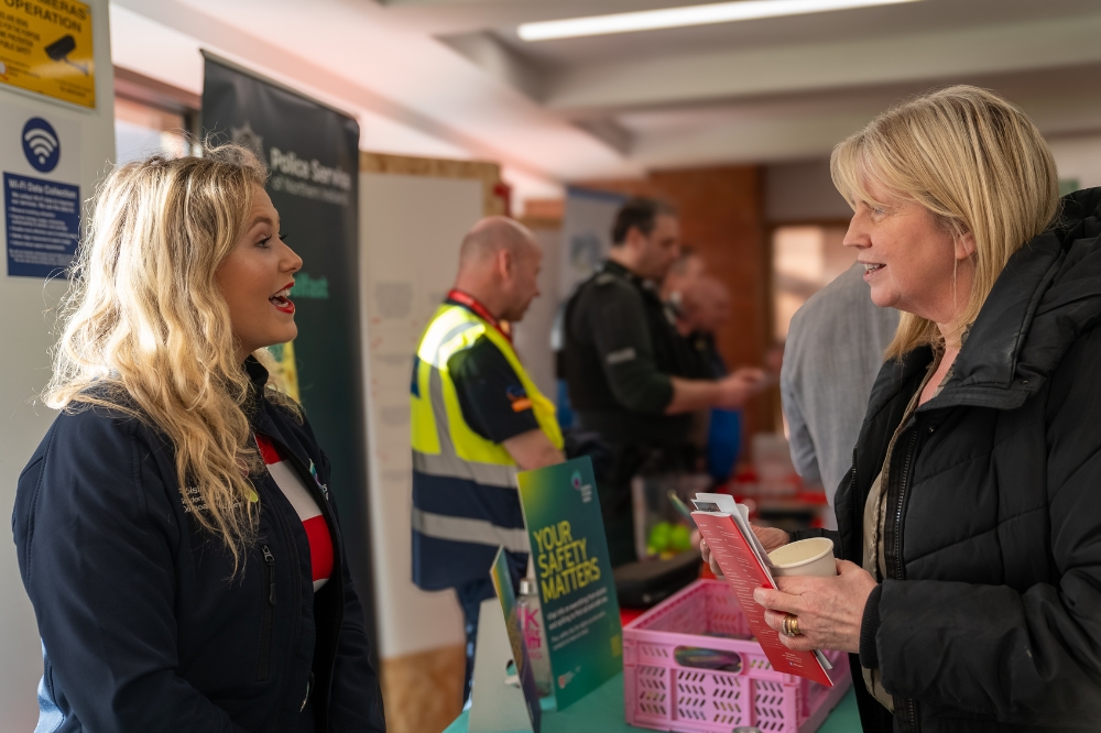 Two people talk at a campus information stall, with safety leaflets and event displays visible in the background.