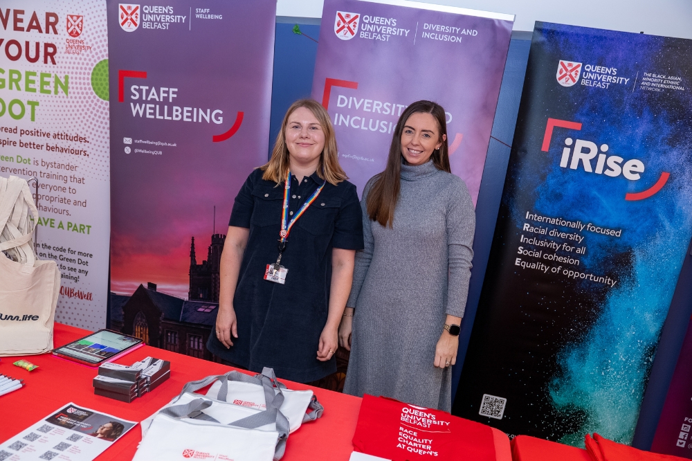Two staff members stand smiling at a Queen’s University Belfast information stall, with Staff Wellbeing, Diversity and Inclusion, and iRise banners behind them