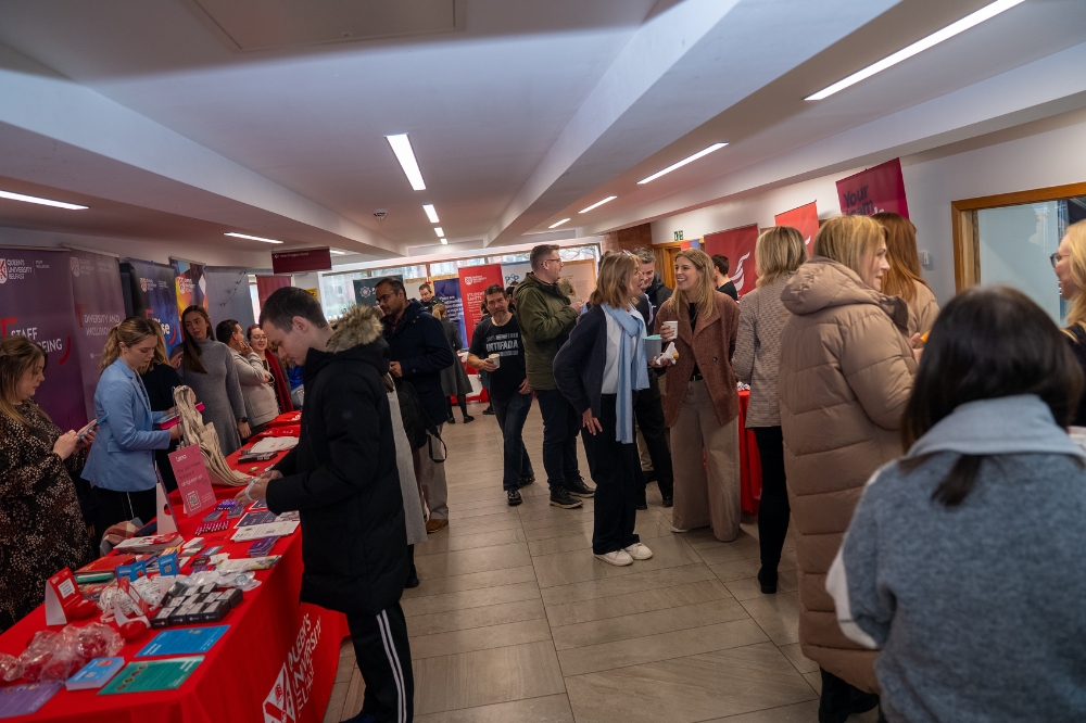 Wide view of a busy indoor campus event with staff and students browsing information stalls and talking with exhibitors along a corridor lined with Queen’s University Belfast banners.