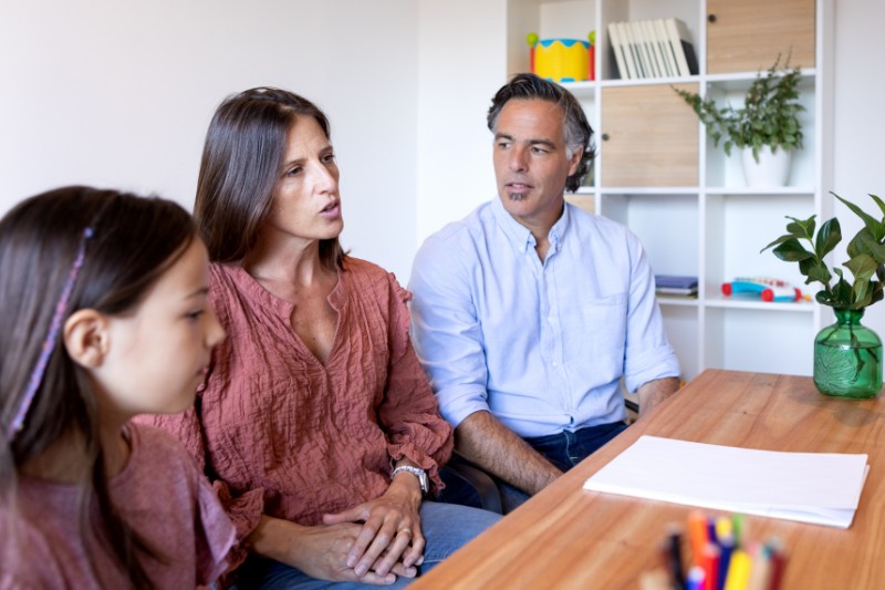 Family (wife, husband/partner and young daughter) in a clean home space. Wife talking to person off-camera, and husband listening and looking on.