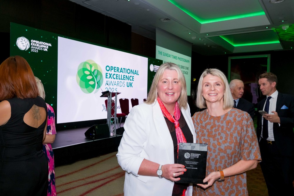 Two women at an awards event holding a trophy, with other event attendees milling around in the background. Background presentation screen shows Operational Excellence Awards UK 2025 logo.