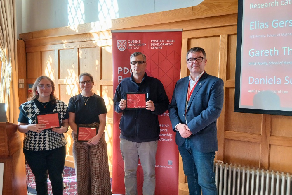 four people, three holding award certificates, at an indoor event in a brightly lit, wood-paneled room
