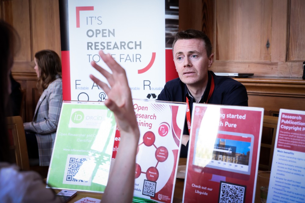 serious-looking man seated and listening to someone chatting to him at a table showing supportive printed materials. Background pop-up stand mentions Open Research.