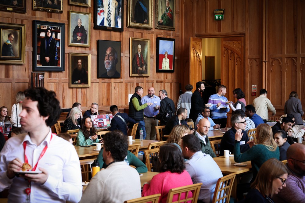 large group of people sitting, standing and enjoying light refreshments in a grand, wood-paneled hall with portraits hanging on the back wall