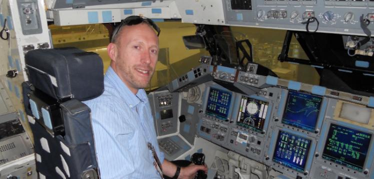 Man wearing blue shirt sitting in cockpit of space shuttle