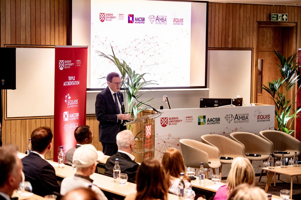 male speaker pictured standing in front of a presentation screen addressing a seated group of attendees in a bright lecture space