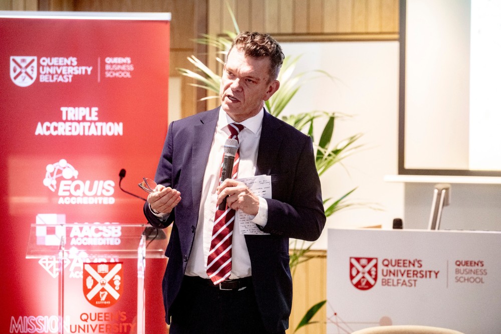 man in business suit holding a notebook and addressing attendees at an event - a pop-up stand and dais are shown in the background