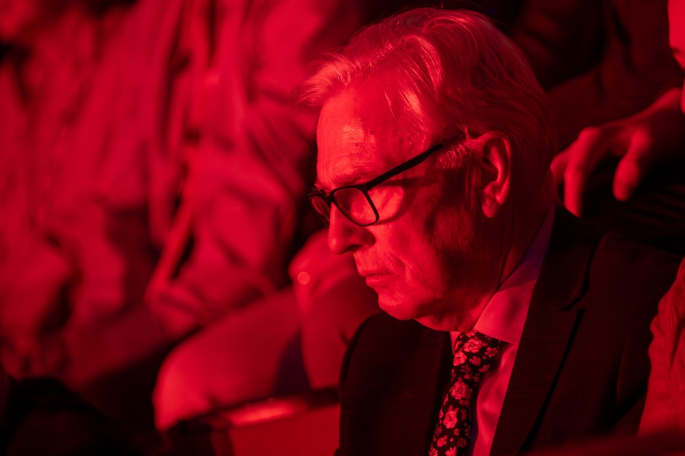 serious looking besuited man seated in a red-lit space watching something off-camera