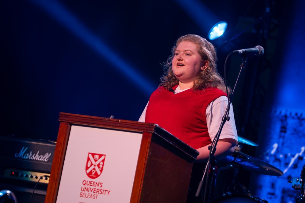 woman wearing a shirt and red vest-jumper speaking at a podium on a stage