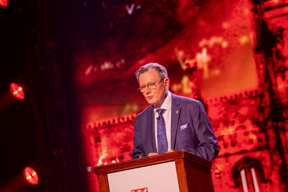 man in suit speaking from a podium against a red presentation screen backdrop