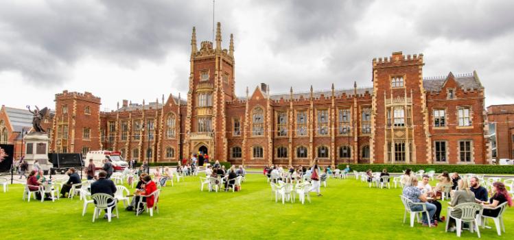 staff and students seated on the Queen's University Belfast front lawn at Queen's Pride Picnic 2024, with food trucks to the side and coloured bunting and balloons in sight