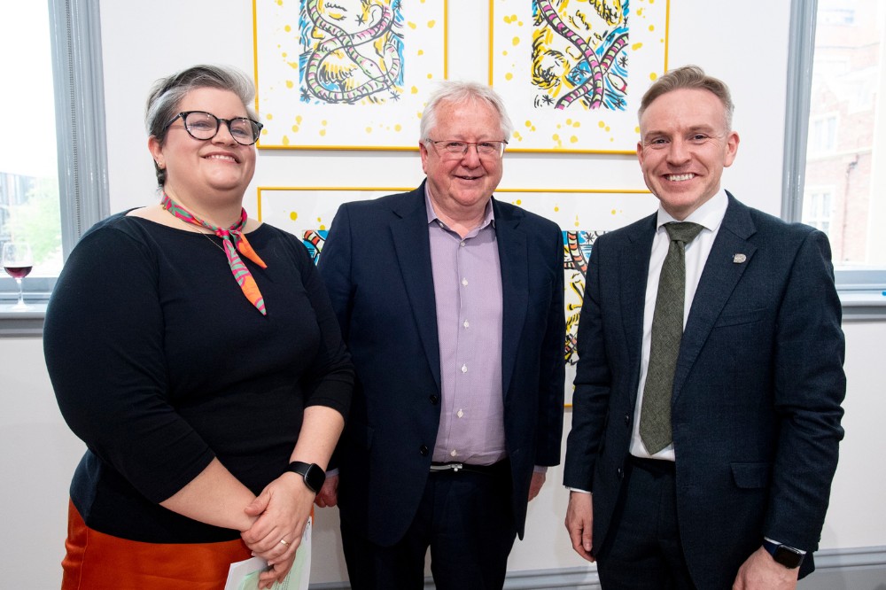 woman and two men smiling for the camera in front of framed artworks on the wall of an an art gallery