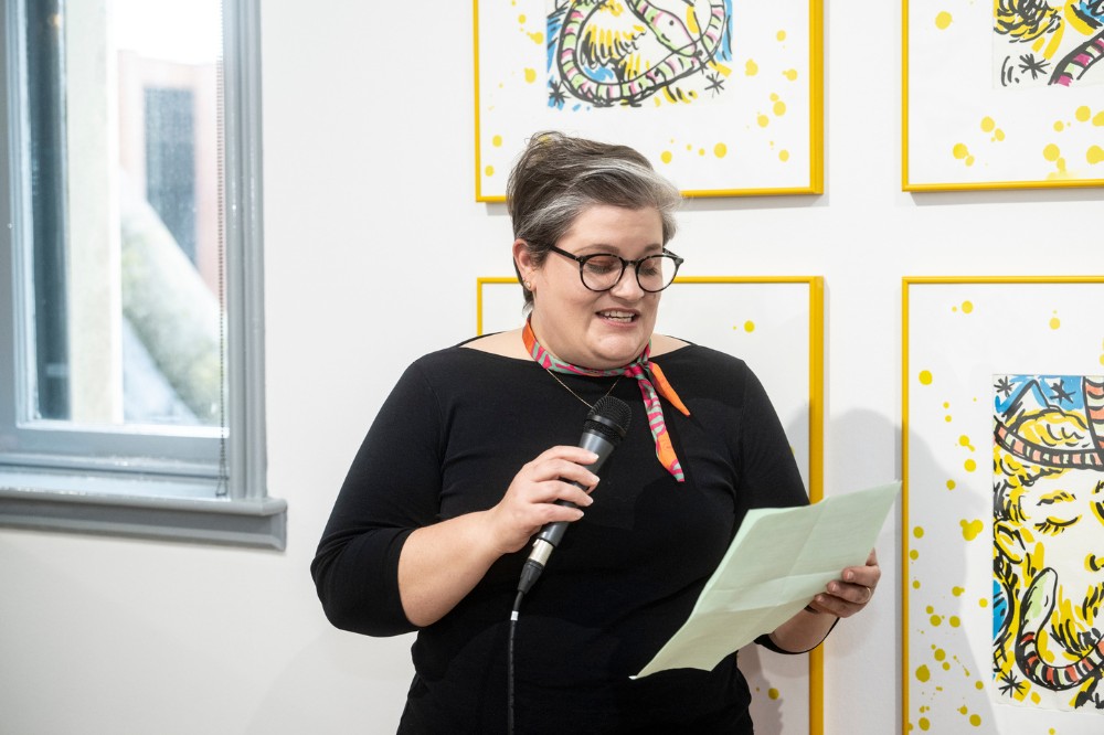 woman holding a microphone reading from a page in an art gallery space