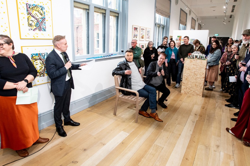 a man in a suit addresses a gathered audience in an art gallery space