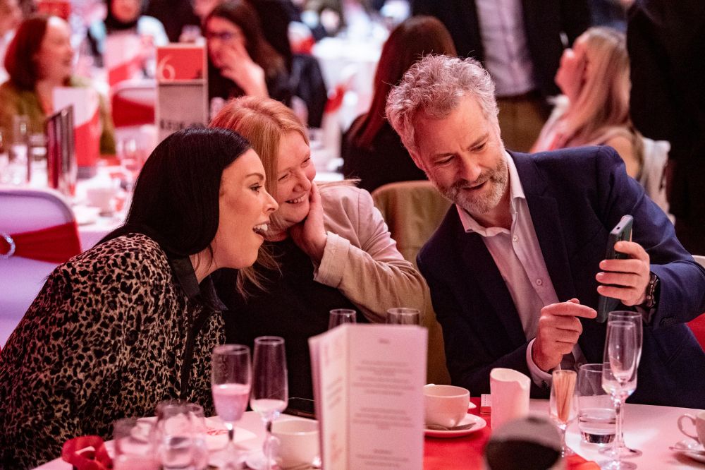 three smiling people seated at a table with man on right showing something on his phone to two women on the left at an indoor event