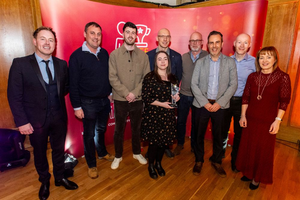 Group of eight men and two women posing to camera at an indoor event. The woman in the centre of the picture is holding a crystal trophy.