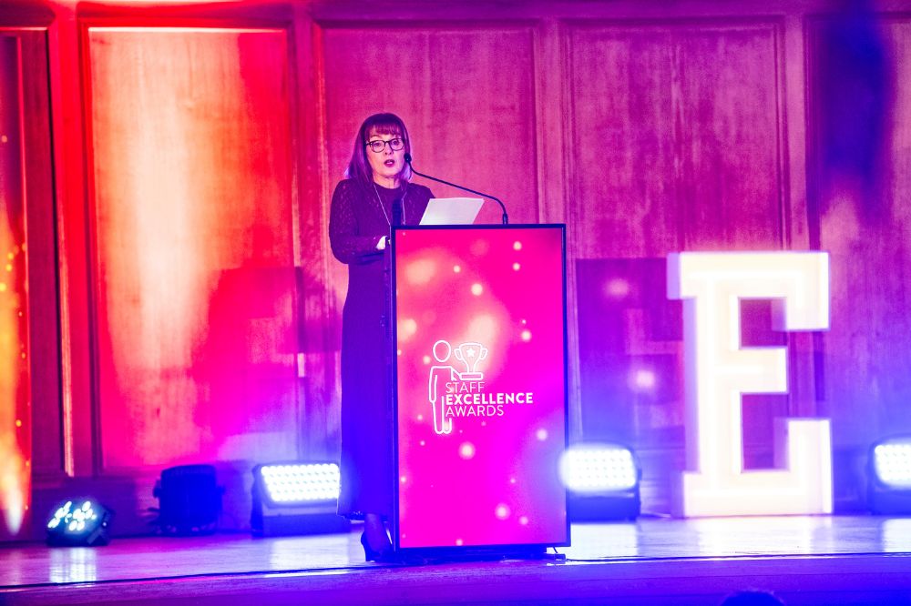 Woman speaking from a podium on a stage with bright light-boxes behind her. The podium façade reads 'Staff Excellence Awards'.