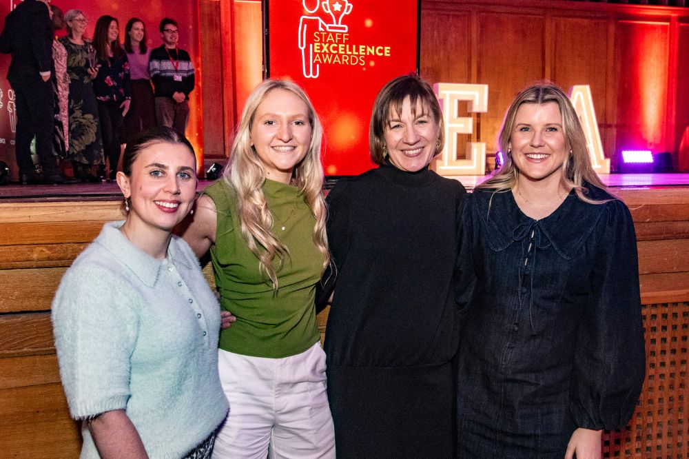four women posing for a photo at an indoor 'Staff Excellence Awards' event, with other people posing for photos in the left background
