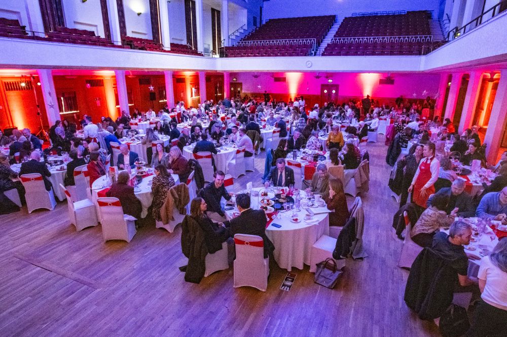 groups of people sitting at dining tables at an indoor event in a large hall with balcony