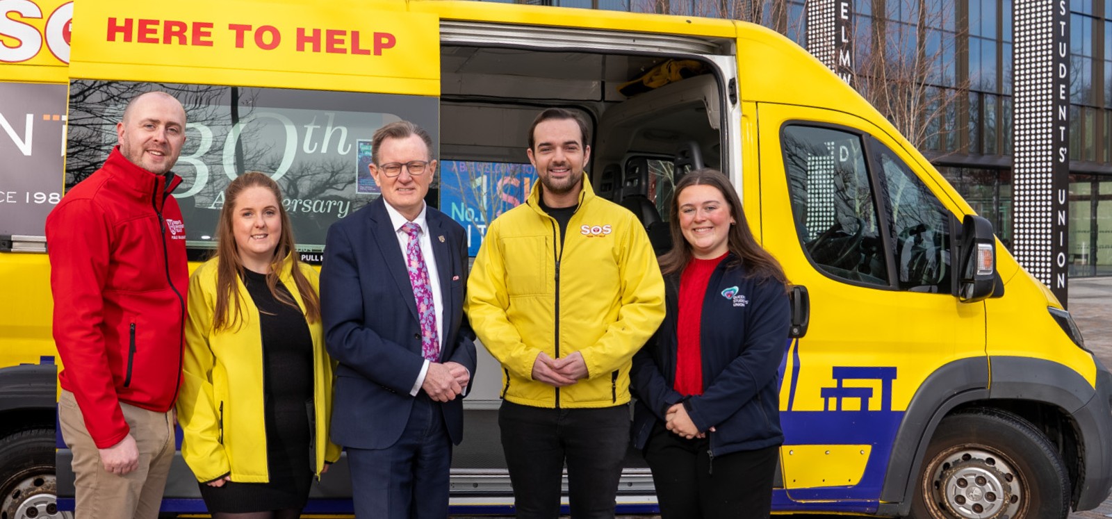 three men and two women in bright business clothing standing in front of a yellow bus