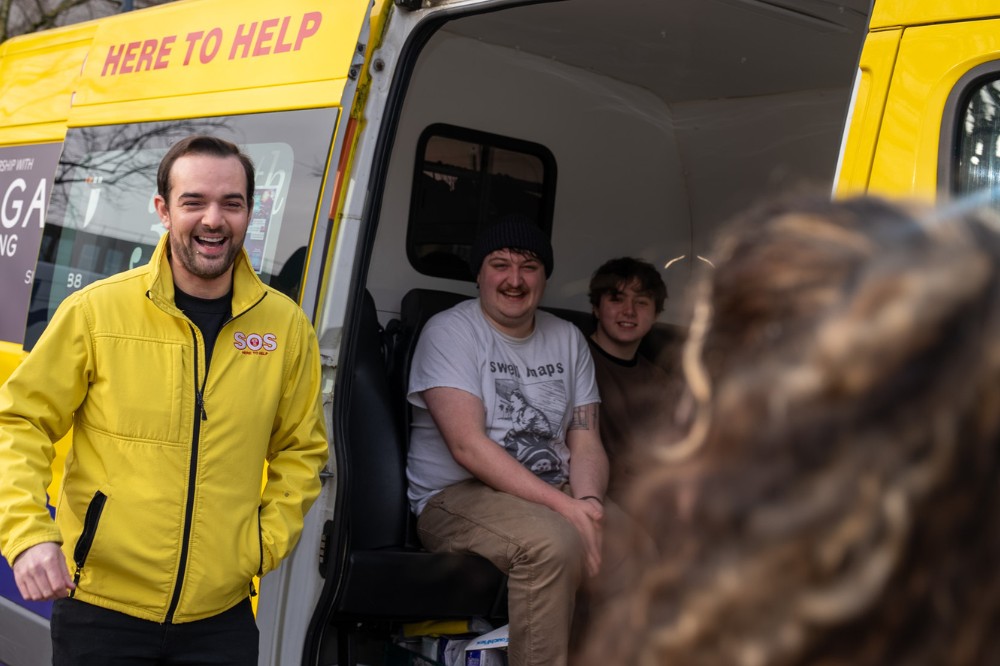 man in yellow jacket sharing a joke and laughing with others beside and seated inside a yellow minibus