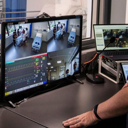 man watching CCTV monitors and electrical equipment behind tinted glass while people perform a simulated scenario in a hospital teaching space