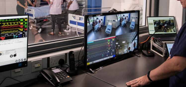 man watching CCTV monitors and electrical equipment behind tinted glass while people perform a simulated scenario in a hospital teaching space