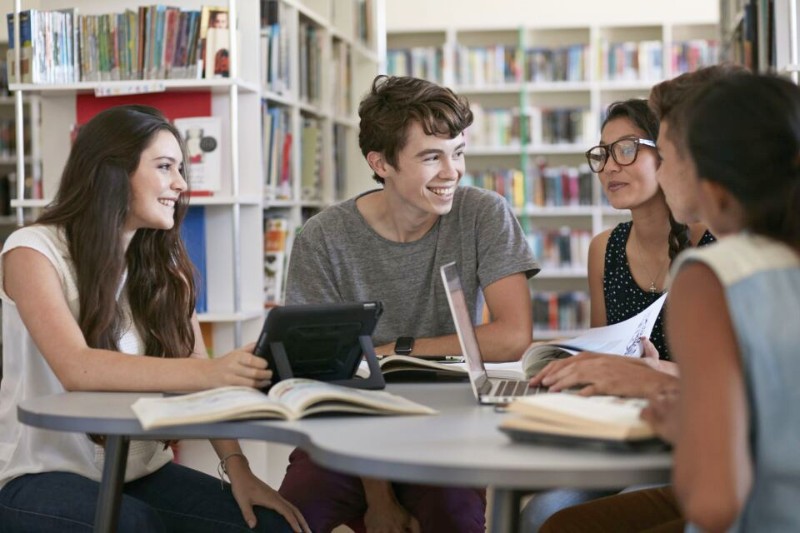 group of four third-level students with laptops and books sitting at a desk and chatting happily