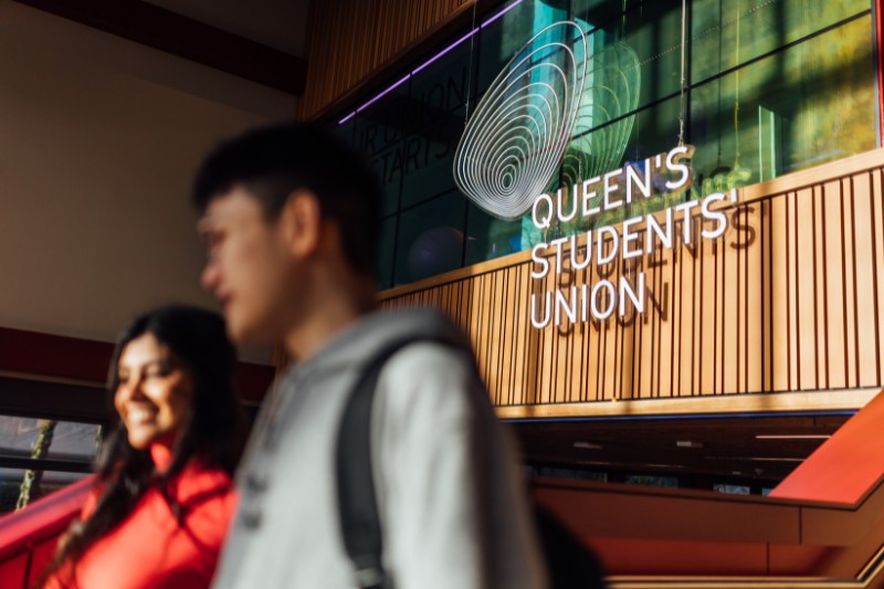 young woman and man, slightly blurred in the foreground, walking past LED lighting reading 'Queen's Students' Union'