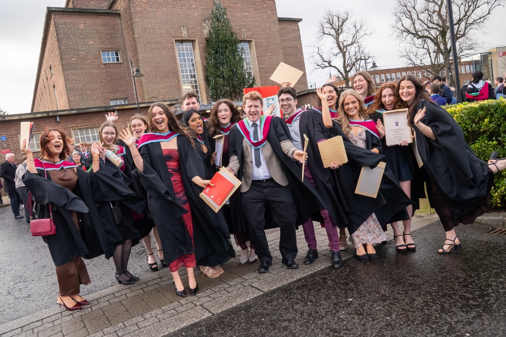 large group of new graduates celebrating their achievement outside a hall with Christmas tree pictured in the background