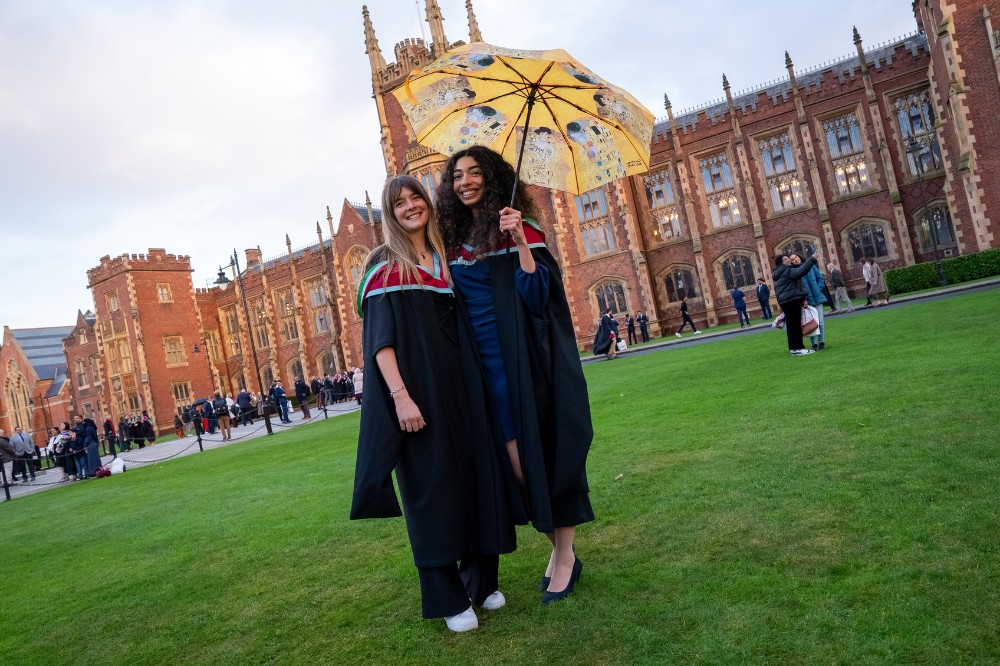 two smiling young women in graduation robes - one holding an umbrella or parasol - standing on the lawn of an old redbrick building