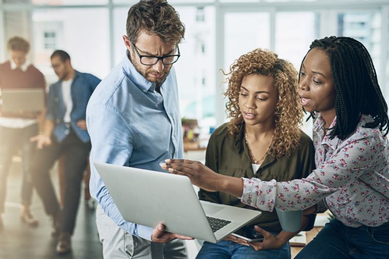 two women offering their thoughts on something a man is showing them on a laptop in a breakout space or workplace environment